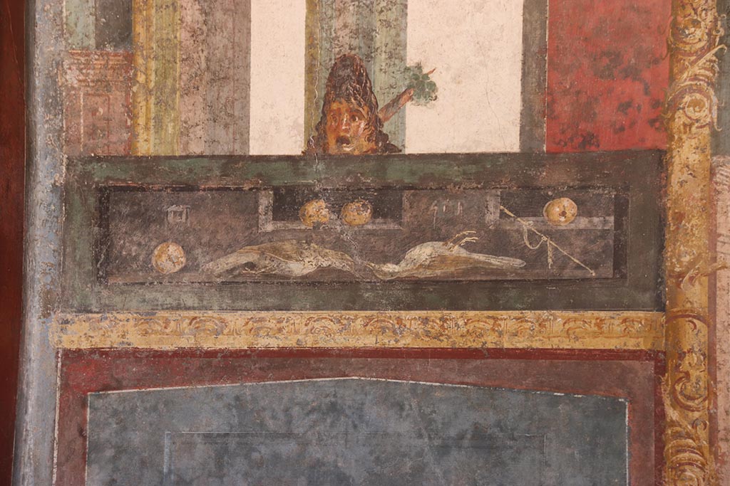 VI.15.1 Pompeii. October 2024.
Detail of panel from east wall at north end, with wall painting of food with mask above. Photo courtesy of Klaus Heese.