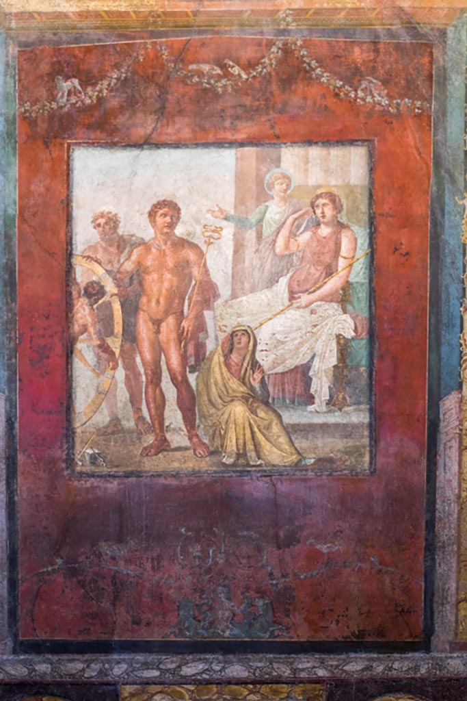 VI.15.1 Pompeii. March 2023.
Central wall painting from east wall of exedra - the torture of Ixion on the wheel.
Photo courtesy of Johannes Eber.
Ixion has already been attached to the wheel by Hephaestus.
Hermes/Mercury stands to the right with his caduceus, helmet and sandals.
The figure seated on the floor is perhaps the cloud nymph Nephele or a personification of the underworld.
Hera/Juno is seated on her throne with Iris standing to her left, gesticulating.
See Carratelli, G. P., 1990-2003. Pompei: Pitture e Mosaici: Vol. V. Roma: Istituto della enciclopedia italiana, p.538.