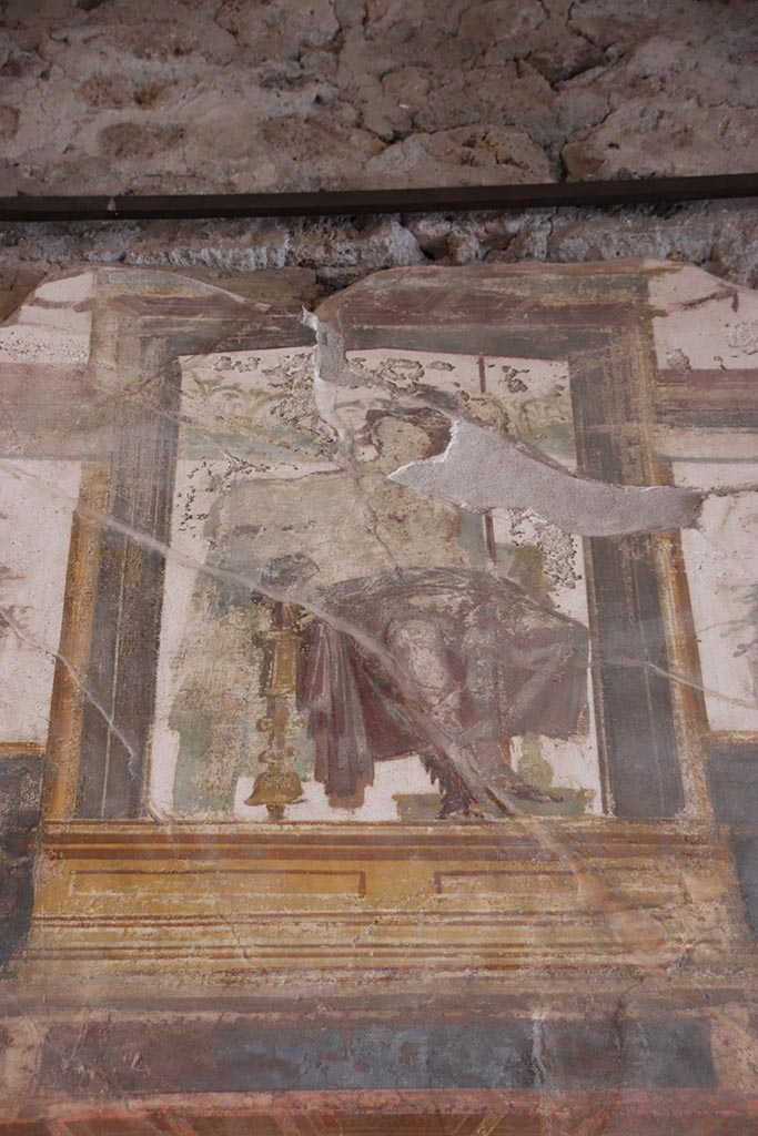 VI.15.1 Pompeii. October 2024.
Upper south wall, painted seated central figure from above central panel. Photo courtesy of Klaus Heese.
In the upper centre of the south wall, damaged, a figure with left shoulder destroyed, and fractures throughout the painting.
Kuivalainen describes –
“Inside an aedicula on a white background, a youth sits on a throne with his legs on a footstool: he leans with his left hand on a long thyrsus and wears high boots and a red cloak on his hips: the yellowish legs of the throne are turned.
Kuivalainen comments –
“The whole south wall is thematically Bacchic, and this young half-naked Bacchus is depicted among other gods in the architecture of the upper zone.”
See Kuivalainen, I., 2021. The Portrayal of Pompeian Bacchus. Commentationes Humanarum Litterarum 140. Helsinki: Finnish Society of Sciences and Letters, (p.100-1, B11).