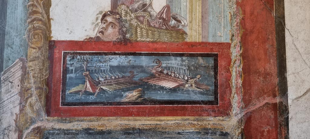 VI.15.1 Pompeii. January 2023.
North wall of exedra with wall painting of naval scene, with basket and mask above. Photo courtesy of Miriam Colomer.