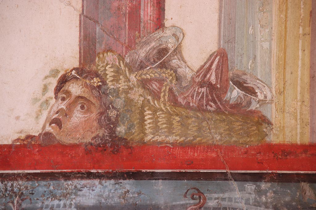 VI.15.1 Pompeii. October 2024.
North wall of exedra with wall painting of detail of basket and mask above the naval scene. Photo courtesy of Klaus Heese.