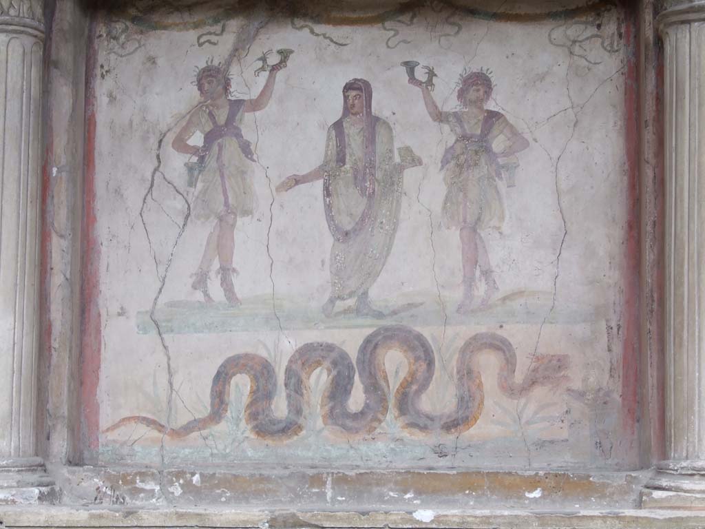 VI.15.1 Pompeii. December 2006. Household lararium in service area.
Painting of serpent, (Agathodemone), garlands and three figures.
The household gods (Lares) are either side of the guardian spirit (genius loci) of the house.
According to Boyce, this was located in VI.15.2.
He described it as one of the largest and finest lararia in Pompeii, consisting of a lararium painting within an aedicula.
The back wall within the aedicula is filled with a lararium painting on a white background.
In the centre stands the Genius, young and unbearded.
On each side of him stands a wreathed Lar, holding rhyton and situla.
The lower end of each rhyton has the form of a forepart of a goat.
In the lower zone of the same panel a crested and bearded serpent with brown back and yellow belly glides through plants towards the altar.
This painted square altar is shown at the right edge of the panel.
On the altar are an egg and some fruits.
Across the top of the panel are three garlands.
See Boyce G. K., 1937. Corpus of the Lararia of Pompeii. Rome: MAAR 14. (p.54, no.211, and Pl.30,2)
See Giacobello, F., 2008. Larari Pompeiani: Iconografia e culto dei Lari in ambito domestico. Milano: LED Edizioni. (p.180-81)
See Fröhlich, T., 1991. Lararien und Fassadenbilder in den Vesuvstädten. Mainz: von Zabern. (p.279, L70).