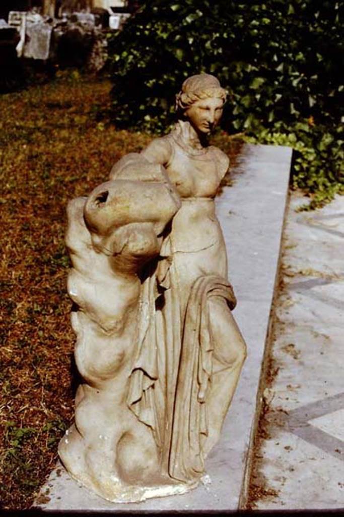 VI.15.5. Pompeii. 1971. Marble fountain statuette of a standing nymph. Photo by Stanley A. Jashemski.
Source: The Wilhelmina and Stanley A. Jashemski archive in the University of Maryland Library, Special Collections (See collection page) and made available under the Creative Commons Attribution-Non Commercial License v.4. See Licence and use details.
J71f0293
According to Wilhelmina, “found inside the niche was a white marble puteal, covered with a grey marble plate on which stood a marble fountain statuette of a standing nymph (0.54m high, Naples Archaeological Museum inv.no: 124 841) leaning with her left arm on a leather bag placed on top of a trunk of a tree. 
The statuette still showed traces of colour. There were various statuettes nearby.” 
See Jashemski, W. F., 1993. The Gardens of Pompeii, Volume II: Appendices. New York: Caratzas. (p.156)