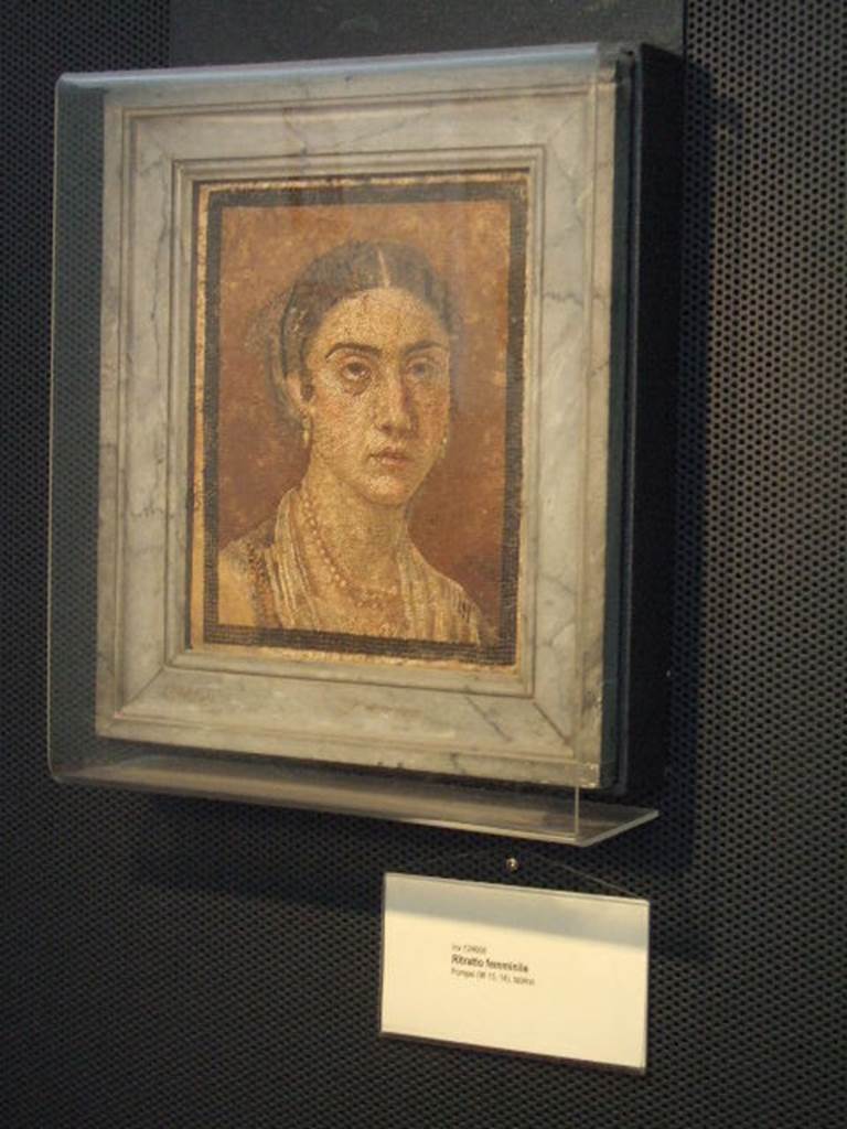 Found in Tablinum of VI.15.14, according to card at Naples Archaeological Museum.  Now in Naples Archaeological Museum, inventory number 124666.  The mosaic shows a young woman but with a matronly aspect, with black wavy hair and pearl earrings mounted in gold.
She is also wearing a pearl necklace, and a dark robe with gold embroidery.
According to Della Corte, the mosaic formed the centre of a floor of a cubiculum situated to the south of the impluvium. (note 1 = N.S. 1898, p.173). See Della Corte, M., 1965.  Case ed Abitanti di Pompei. Napoli: Fausto Fiorentino. (p.63)
According to NdS, the mosaic was found on 6th April 1898, in the centre of a floor of a cubiculum. It was contained in a piece of travertine, framed by a strip of black mosaic and made of very small tesserae, finely executed. The only damage was to the lower left corner of the mosaic, at the woman’s right shoulder. See Notizie degli Scavi, April 1898 (p.173)