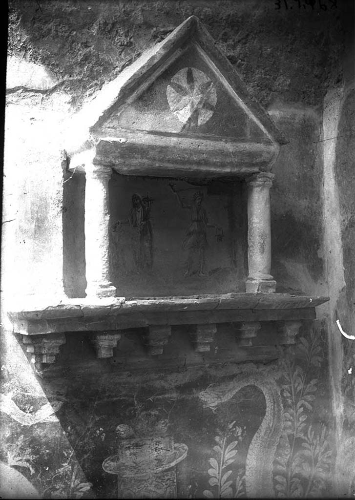 VI.15.23 Pompeii. Lararium on the west wall, in the north-west corner of the kitchen.
DAIR 31.2468. Photo © Deutsches Archäologisches Institut, Abteilung Rom, Arkiv. 
According to Boyce, the whole was painted red except for a star on a white ground in the centre of the tympanum.
The curved back wall of the niche was painted blue, and upon it was the lararium painting.
On each side was a Lar, between them stood the Genius.
On the wall on each side of the aedicula was painted a garland and a flying bird.
Two large yellow serpents were painted on the wall beneath the aedicula and extended over onto the north wall on the one side.
On the other side, extending to the adjacent side of the hearth.  
The serpent on the right had the larger body, a crest and a beard, the one on the left was smaller with no crest or beard.
They were on either side of a cylindrical altar painted in imitation of yellow marble beneath the aedicula.
The altar was furnished with two eggs and fruit.
See Boyce G. K., 1937. Corpus of the Lararia of Pompeii. Rome: MAAR 14.  (p.56, no.219, and Pl.15, 1 and 2)
See Giacobello, F., 2008. Larari Pompeiani: Iconografia e culto dei Lari in ambito domestico. Milano: LED Edizioni. (p.183)