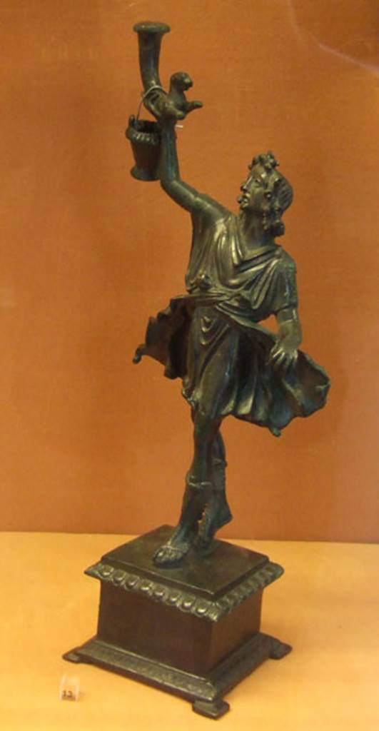 Bronze statuette of a Lar, found in Lararium of VI.16.7. 
This was one of two Lares that were found on the lower step of the lararium.
The Lar holds a rhyton which has at its lower end the front half of a panther.
The bucket would normally be in the Lar’s left hand.
Now in Naples Archaeological Museum. Inventory number 133328.
See Notizie degli Scavi di Antichità, 1907, p. 567, fig. 17.