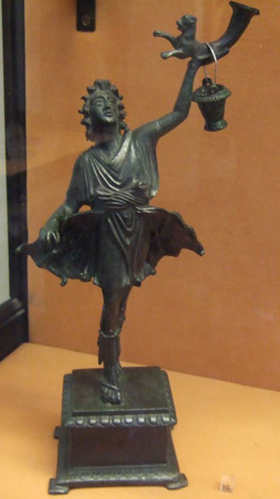 Bronze statuette of a Lar, found in Lararium of VI.16.7. 
This was one of two Lares that were found on the lower step of the lararium.
The Lar holds a rhyton which has at its lower end the front half of a panther.
The bucket would normally be in the Lar’s right hand.
Now in Naples Archaeological Museum. Inventory number 133327.
See Notizie degli Scavi di Antichità, 1907, p. 567, fig. 17.