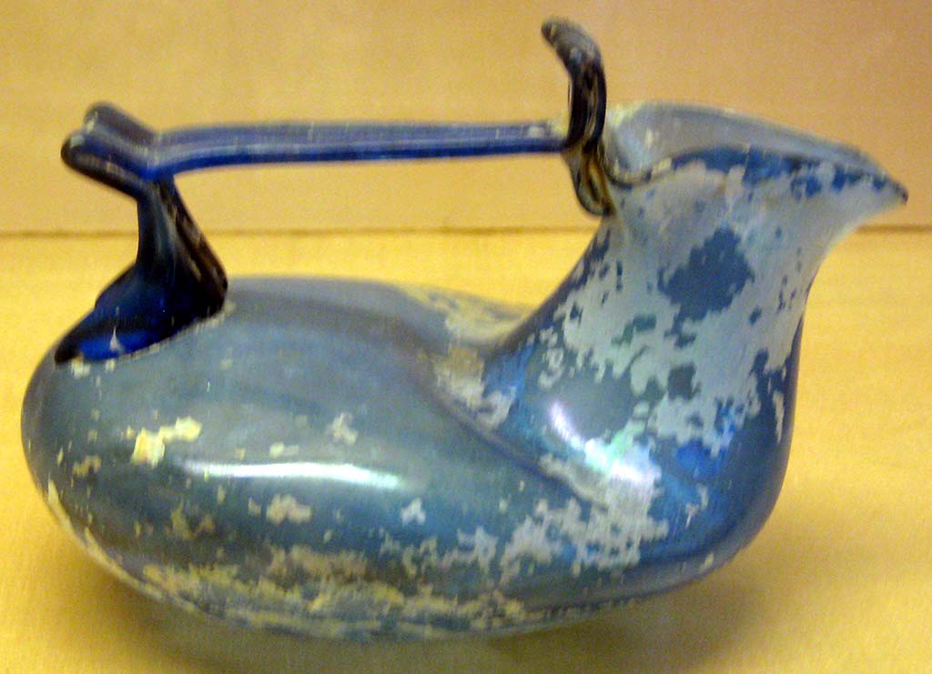 VI.16.10 Pompeii. Deep blue glass askos with encrustation found here. 
It has a trilobate mouth, rigid handle with flat ribbon with rounded apex, lower bifurcated graft, 
Now in Naples Archaeological Museum. Inventory number 133275.