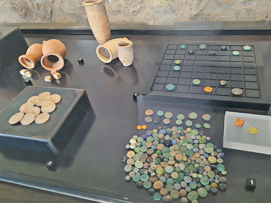 VI.16.27 Pompeii. March 2024. No.3 above, gaming counters made of worked bone. Photo courtesy of Giuseppe Ciaramella.
On display in exhibition in Palaestra entitled – “L’altra Pompei, vite comuni all’ombra del Vesuvio”. March 2024.
No.1 – Gaming counters made of glass paste, die and tesserae made of bone, from Porta Stabia Necropolis.
No.2 – Worked bone dice, from V.4.13, Pompeii.
No.3 – Gaming counters made of worked bone, from VI.16.26/27, Pompeii.
No.5 – Pottery dice shakers, from II.1.6, Pompeii.
No.6 – Reproduction of a gaming table.