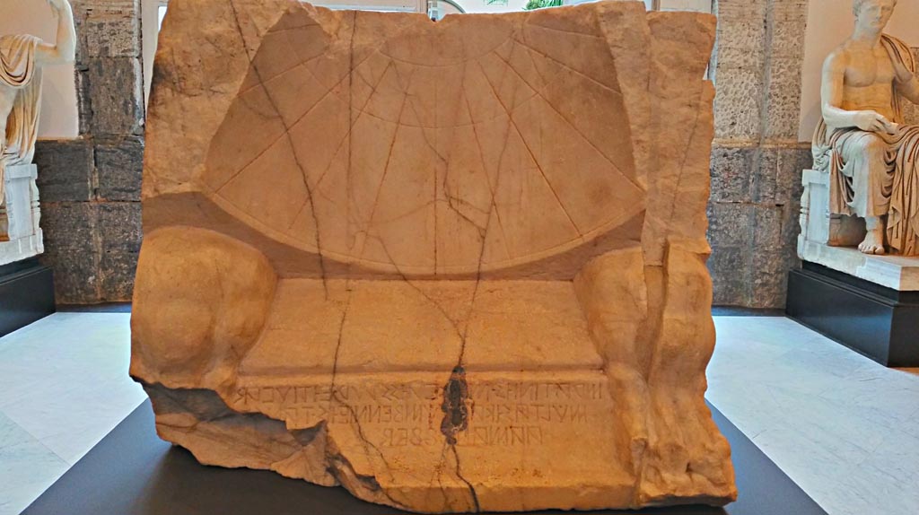 VII.1.8 Pompeii. November 2018. 
Marble sundial with Oscan inscription, found 23rd September 1854. Photo courtesy of Giuseppe Ciaramella.
An inscription in Oscan reveals that the quaestor Maras Atinius commissioned the construction of the sundial:
“Maras Artinius (son of) Maras, quaestor, with the money from the fines, by resolution of the assembly, had (this) made”.
Solar clocks were one of the many contributions of the Greeks to the Roman world, and this refined Pompeian specimen bears witness to the Hellenistic taste to which the Terme Stabiane (a modern name referring to their position along the Via Stabiana) conformed. The Terme Stabiane replaced an earlier, much less sophisticated facility in the 2nd century BC.
Discovered at Pompeii on 23 September 1854 in the Stabian Baths, it was used to regulate the turns for entering the complex.
The dial is a portion of a right circular cone, bounded on the upper side by a hyperbola.
The clock has three circular day curves that are parallel to each other.
The day curves are the lines cast by the shadow of the gnomon over the course of a whole day. The arcs of the solstices and equinoxes are each divided into 12 equal parts to plot the eleven hour lines, obtained by joining the corresponding partitions of the three day curves.
The hours marked are “unequal hours”.
Now in Naples Archaeological Museum. Inventory number 2541.