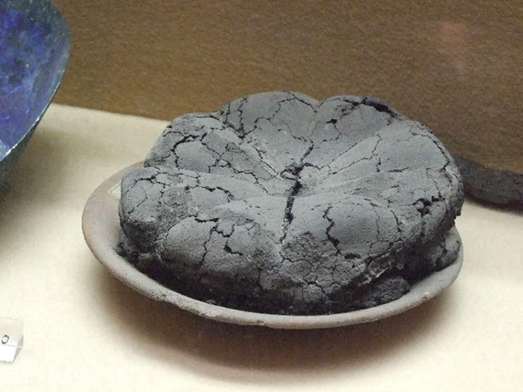 VII.1.36 Pompeii. One of 81 loaves found in bakery ready for sale. Now in Boscoreale Antiquarium. According to Della Corte, the bakery was one of the most complete found up until then in the excavated city, from the tub for the working of the grain, the stall for the shelter of the beasts that worked the four machines, the workshop for the proving of the bread, and the shop, which opened onto the via at number 37, for the sale of the same bread. Precisely in this oven of the bakery, found closed with a door of iron, were 81 carbonised loaves of bread, put there to cook minutes before the catastrophe.
See Della Corte, M., 1965.  Case ed Abitanti di Pompei. Napoli: Fausto Fiorentino. (p.189-90