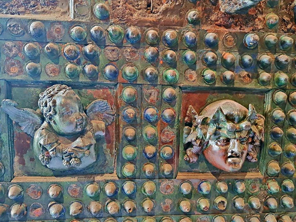 VII.2.18 Pompeii. October 2023. 
Arca/strong box, detail front of box – 
(on left) winged cupid carrying a garland of vine leaves and grapes around the neck, and (on right) a mask with long hair crowned with ivy.
Photo courtesy of Giuseppe Ciaramella.