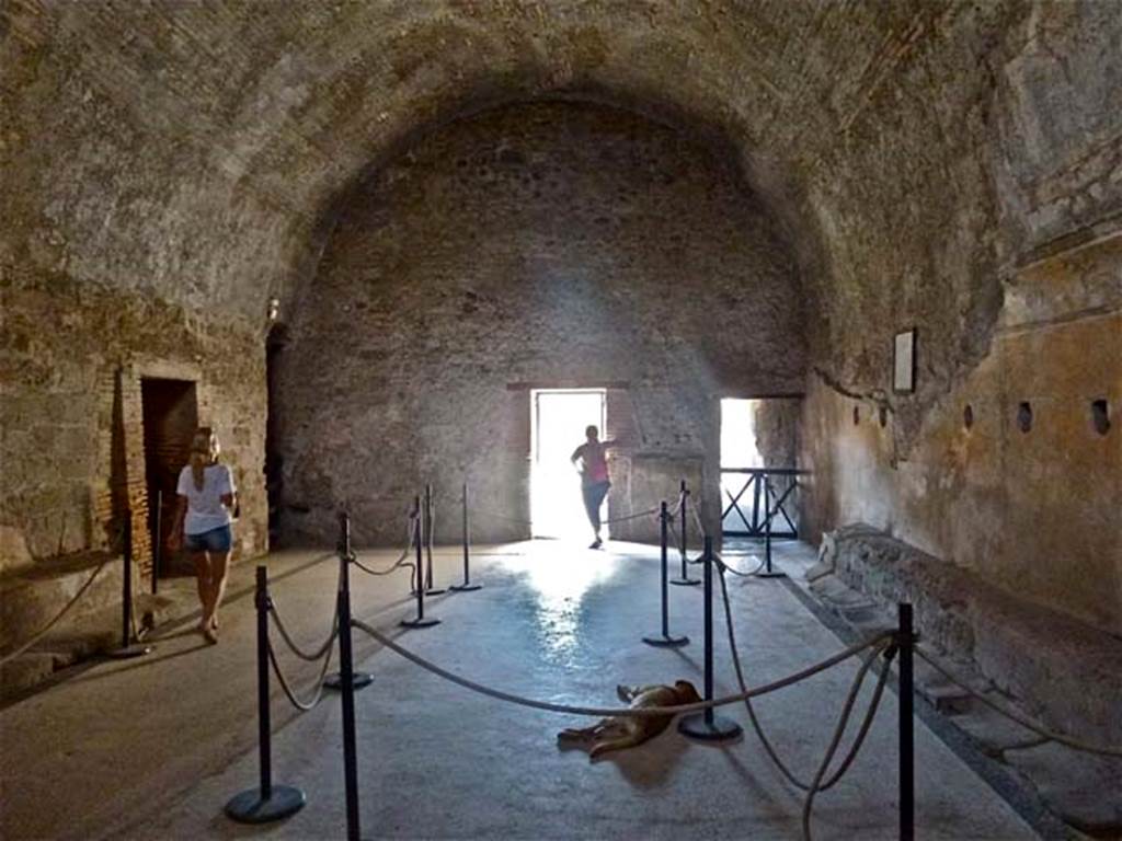 VII.5.24 Pompeii. June 2012. Men’s baths apodyterium or changing room (14), looking north. 
Photo courtesy of Michael Binns.
According to Fiorelli –
The changing room (14), other than the doorway from the fauces (corridor 16, on right above), had another five doorways around it. 
On the right of the corridor 16, (centre, above) was a small room where the oils and ointments were stored.
On the left of the photo above (west) was the doorway to the long corridor leading to the boiler area.
On the left (behind the figure) was also the third doorway which led into the tepidarium (37).
The fourth doorway (in the south-west corner) led into the frigidarium (19)
The fifth doorway (in the south-east corner) led into corridor 13, leading towards the garden/courtyard area, which had two secondary entrances or exits towards the east and west sides of the Insula.
See Pappalardo, U., 2001. La Descrizione di Pompei per Giuseppe Fiorelli (1875). Napoli: Massa Editore. (p.94-95).