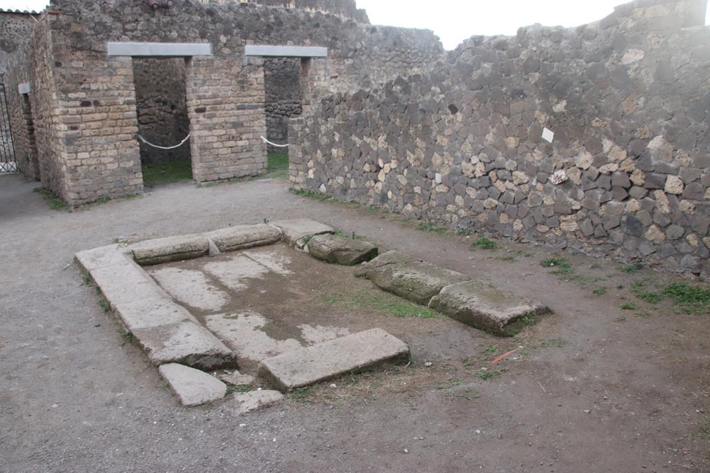 VII.7.2 Pompeii. October 2024.
Looking south across impluvium “g” made of slabs of tufa, from south side of tablinum “k”. 
The doorway to room “d” is on the south side, with doorway to room “i” visible behind the wall.  Photo courtesy of Klaus Heese.