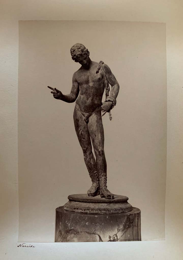 VII.12.17.21 Pompeii. Photograph by M. Amodio, from an album dated April 1878.
Statuette of Narcissus also sometimes described as Dionysus or Pan. Photo courtesy of Rick Bauer.
According to Kuivalainen, this statue (now in Naples Archaeological Museum, inv. no: 5003), had fallen from the upper floor.
“The condition is good, but the original base has been replaced, and the panther is lost.”
He comments – 
“The male’s apparel and the position of the right-hand fingers identifies the bronze as Bacchus dangling a bunch of grapes above a panther.
The iconographic formula is also known from wall paintings in Pompeii.”
See Kuivalainen, I., 2021. The Portrayal of Pompeian Bacchus. Commentationes Humanarum Litterarum 140. Helsinki: Finnish Society of Sciences and Letters, (p.219, H14.)
.