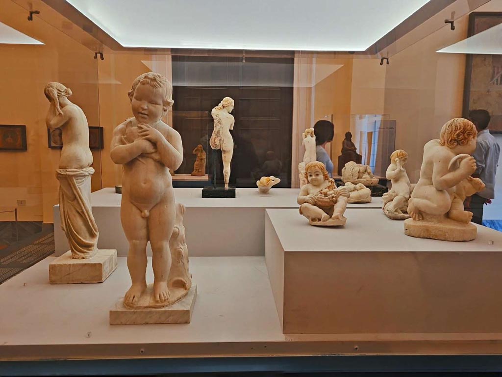 VII.12.23 Pompeii. October 2023. Group of statuettes, on right, found in the garden area.
On the left is a marble statuette of a child caressing a dove, from vesuvian area, but location unknown, (inv. 6114).
Photo courtesy of Giuseppe Ciaramella. 
On display in “L’altra MANN” exhibition, October 2023, at Naples Archaeological Museum.