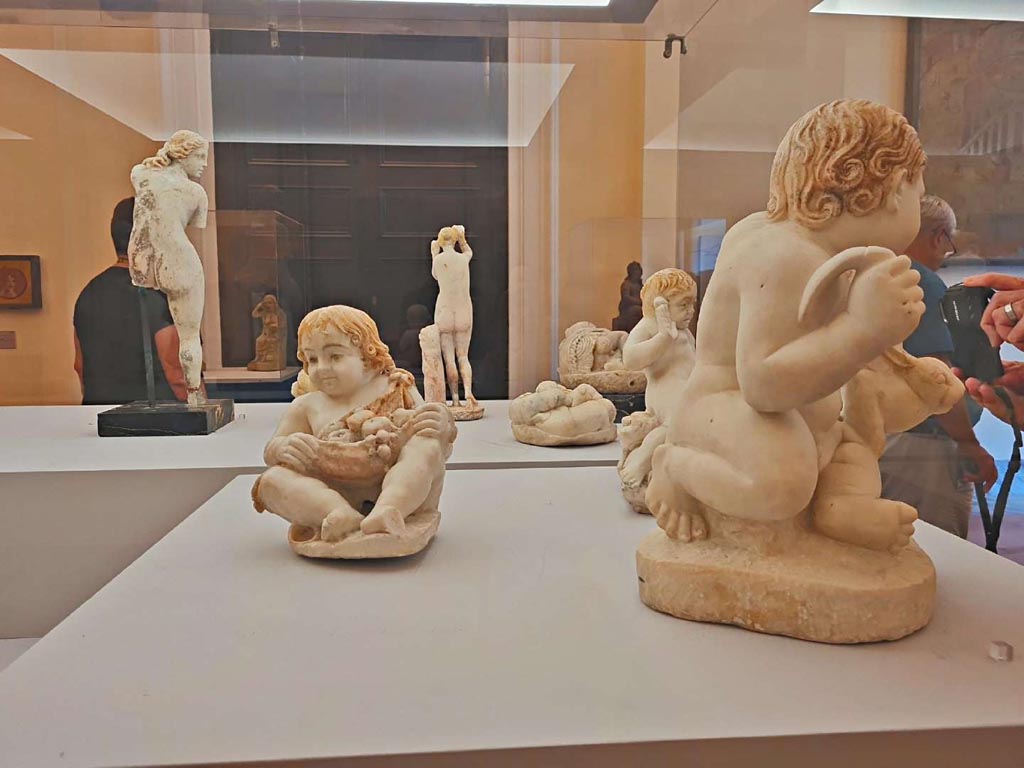 VII.12.23 Pompeii. October 2023. Group of statuettes, found in the garden area.
On the left – (10) Fountain figure of child holding fruits in his mantle, (inv. 6500).
On the right – (12) Fountain figure of a child holding up a hare, (inv. 6533).
At the rear – (11) Fountain figure of a child frightened by a toad, (inv. 6537).
Photo courtesy of Giuseppe Ciaramella. 
On display in “L’altra MANN” exhibition, October 2023, at Naples Archaeological Museum.