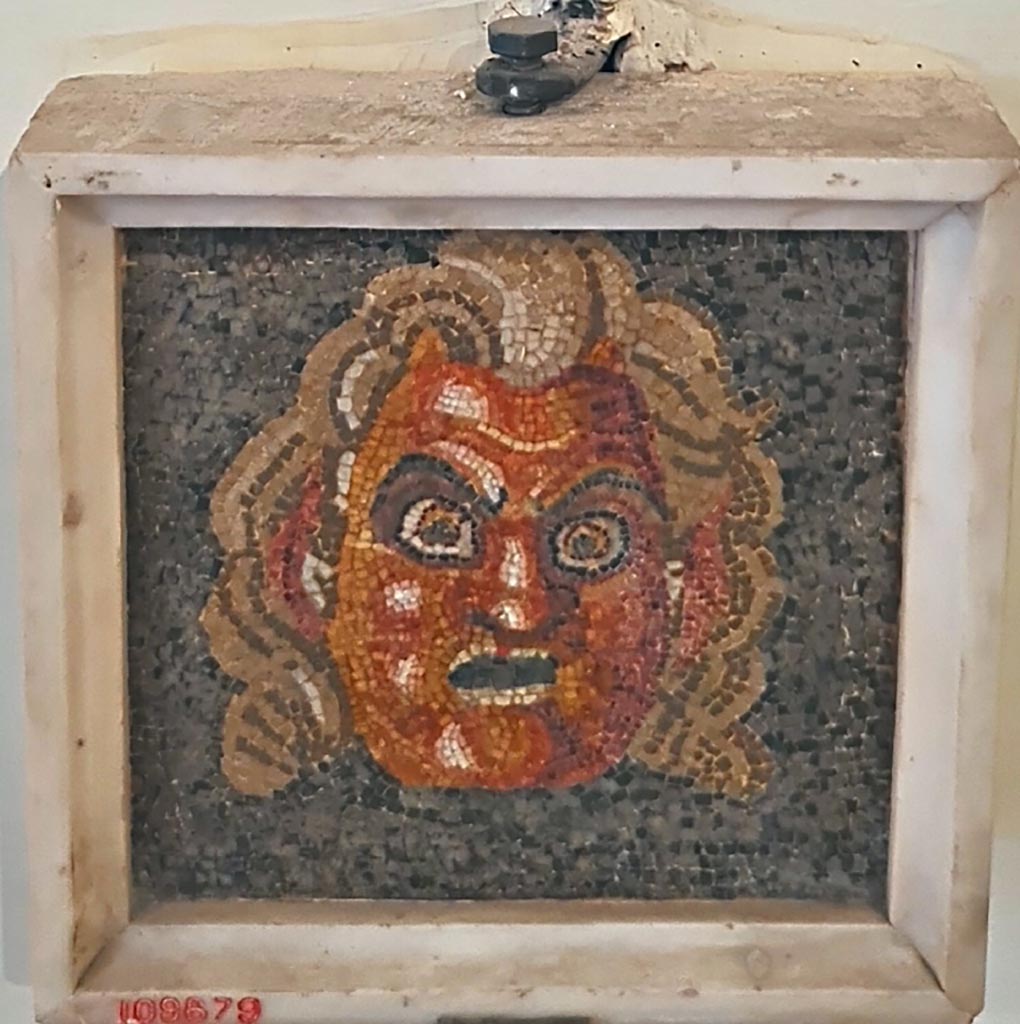 Mosaic mask found at VII.13.23 on 17th November 1821. 
Now in Naples Archaeological Museum. Inventory number 109679. 
According to the Giornale, in this location were found two beautiful masks in mosaic embedded above two stones of marble.
See Fiorelli G., 1864. Pompeianarum antiquitatum historia, Vol. 3: Naples, p. 30, dated 17th November 1821.
According to the information card –
Theatrical mask.
Fragment of a mosaic pavement in opus vermiculatum made from polychrome tesserae on a black background, depicting a theatrical mask of Pan.  (Early Ist century BC.)
(Maschera teatrale.
Frammento di mosaico pavimentale in opus vermiculatum di tessere policrome su fondo nero, raffigurante una maschera teatrale di Pan. 
Inizi del I secolo a.C.)
Photo courtesy of Giuseppe Ciaramella. September 2024.