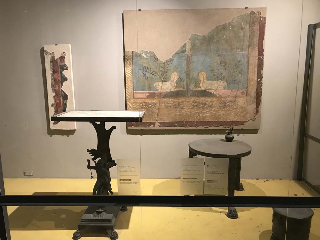 VII.16.17-22 Pompeii. April 2019. On display in Antiquarium, from House of Fabius Rufus. 
Bronze table with cupid riding a dolphin and marble top. PAP inventory number 13371.
Bronze table with lion's feet. PAP inventory number 13108.
On table is a bronze pitcher with face of a follower of Dionysus. PAP inventory number 14072.
Fragment of fresco on the wall at the rear, from VI.17.42, Pompeii. 
Fragment of the wall of the summer triclinium 31 decorated with a pair of facing Sphynxes within a lush garden.
Parco Archeologico di Pompei, inventory number 87228.
Photo courtesy of Rick Bauer.