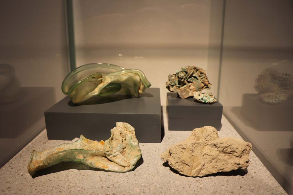 VIII.1.4 Pompeii. February 2021. Items on display in antiquarium.
Upper and lower left, Glass ointment jar and bowl distorted by the eruption, from I.7.11/18.
Upper right, bowl containing nails distorted by the eruption, from VI.16.9.
Impression of an Ilex (holm oak) leaf, lower right, from Villa San Marco.
Photo courtesy of Fabien Bièvre-Perrin (CC BY-NC-SA).