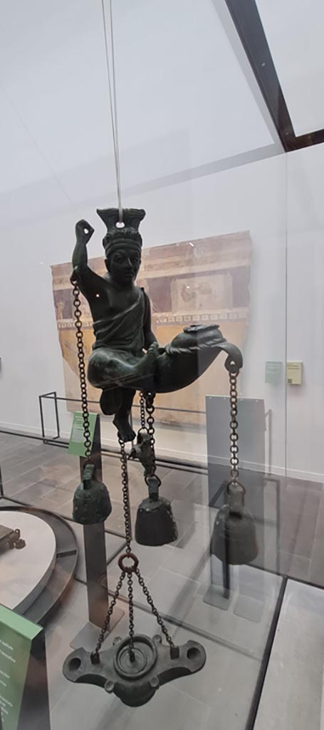 VIII.1.4 Pompeii. April 2022. Tintinnabulum.
Bronze lamp with statuette of a satyr with a phallus from which small bells hang, found in I.6.3.  
Photo courtesy of Giuseppe Ciaramella.