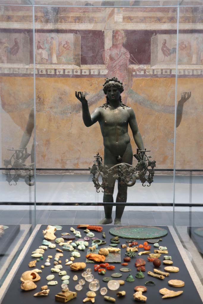 VIII.1.4 Pompeii. February 2021. 
Looking across amulets and gems found in V.3, House with Garden, towards bronze Ephebus lamp from VII.16.19/22. 
At the rear, is part of the painted wall from the oecus/triclinium of I.6.2.
Photo courtesy of Fabien Bièvre-Perrin (CC BY-NC-SA).