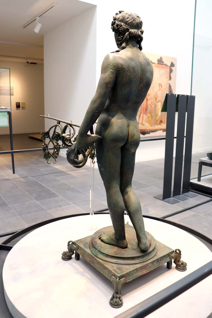 VIII.1.4 Pompeii. February 2021. 
Detail of bronze Ephebus as an oil lamp holder, found in dining room of VII.16.19/22.
Visible on the wall to the right is a wall painting from VI.17.42, House of the Golden Bracelet.
Photo courtesy of Fabien Bièvre-Perrin (CC BY-NC-SA).