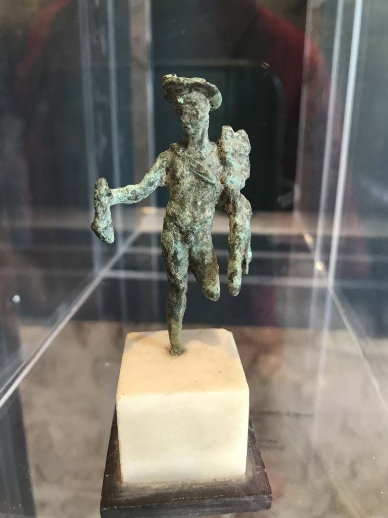 VIII.2.1 Pompeii. April 2019. Bronze statuette of Mercury carrying a money bag.
Found in the area to the south of the House of Championnet.
Pompeii Inventory number 6331. Photo courtesy of Rick Bauer.
According to Zanella -
This statuette was found in Room B, the triclinium on the lower floor of VIII.2.3-5 (or VIII.2.14-16) on 9th February 1937.
9/2/1937
In room B an imprint of a box had been removed at a distance of m. ,0,70 from the bed frame, this imprint had been found to be m.0.68 x 0.50 x 0.22 including a shelf in the front m.0.50 x 0.18 x 0.16.
In removing the last clod of ash with the partial imprint of the triclinium couch, the following was found: Bronze: statue representing Mercury standing missing a (leg) with the pouch and caduceus; it was fragmented and missing its left leg from the knee down and the right foot. 
Alt m.0.068 (Pompeii inv. 6331)
(Nel ambiente B si era rimosso un’impronta di cassa distante m. ,0,70 dal telaio del letto, detta impronta era risultata di m.0.68 x 0.50 x 0.22 compreso un ripiano nella parte anteriore m.0.50 x 0.18 x 0.16.
Nel rimuovere l’ultima zolla di cenere con l’impronta parziale del letto tricliniare si era rinvenuto: bronzo: statuina rappresentante Mercurio stante mancante di un (*) col marsupio e caducco; era frammentata e mancante della gamba sinistra dal ginocchio in giù e del piede destro. Alt m.0.068 (Pompei inv. 6331))
See Zanella S., 2019. La caccia fu buona: Pour une histoire des fouilles à Pompéi de Titus à l’Europe. Naples : Centre Jean Bérard, p. 268.