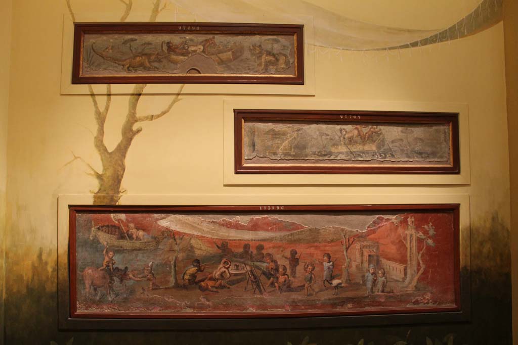 VIII.5.24 Pompeii. March 2014. Display of painting on a wall in Naples Archaeological Museum.
The lowest painting is a Nile banquet scene with pygmies. From a low wall connecting peristyle columns in VIII.5.24.  
The upper paintings are from VII.2.25.
Foto Annette Haug, ERC Grant 681269 DÉCOR.
