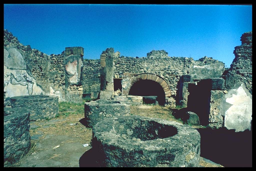 VIII.6.11 Pompeii. Bakery, looking east.
Photographed 1970-79 by Günther Einhorn, picture courtesy of his son Ralf Einhorn.
According to Mau, from the entrance corridor one immediately entered the bakery, where one could see the substructures of four mills, and around them the flooring for the mules.
The walls were painted simply in the last style, with red and yellow compartments.
Near to the north wall was a square stone that was probably used as a table.
A larger table was against the left wall (west), between the two doorways and was supported by two feet of wood, while the other side was supported by two beams fixed into the wall.  The table cannot be currently recognised other than by these two beam holes (height 0.75, distance between them 2.10m) and the two holes in the floor (0.80 distance from the wall) where the wooden feet stood.
A similar table but smaller (1.m) was against the same wall between the doorway to “f” and the front corner (south-west corner).
“a” is the oven, (round, not elliptical, as it seems on the plan); before the oven on the left buried in the ground was a vessel for water, and next on the left was a large dolium.
See Bullettino dell’Instituto di Corrispondenza Archeologica (DAIR), 1884, p.138
(Per la fauce, che ha la porta immediatamente alla strada,s'entra nel pistrinum, ove troviamo le sostruzioni per quattro mulini, e intorno ad esse il selciato per i muli. Le pareti son dipinte semplicemente nell'ultimo stile, con compartimenti rossi e gialli. Addosso al muro N sta una pietra quadra di tufo che serviva probabilmente da tavola. Una tavola più grande stava addosso al muro sinistra (0), fra le due porte, ed era sorretta da due piedi di legno, mentre l'altro lato era con due travi fissato nel muro. Così presentemente la tavola non si riconosce che dai buchi di queste due travi (a. 0,75, distanti fra loro 2,10) e da due buchi corrispondenti nel pavimento, (distanti dal muro 0,80), ove stavano i piedi di legno. Una tavola simile ma più piccola (m. 1) stava addosso alla stessa parete fra la porta di “f" e l'angolo anteriore. – “a” è il forno (tondo, non ellittico, come sembra nella pianta); avanti ad esso sta a sinistra un vaso per acqua murato nel suolo, e accanto a sinistra un gran dolium.)