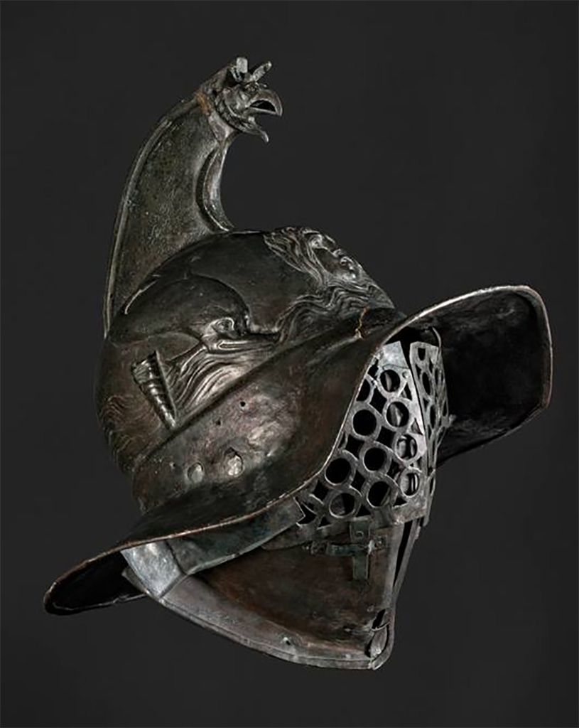 VIII.7.16 Pompeii. Thraex gladiators helmet. 
The crest of this helmet ends in a griffin’s head and feathers could also be attached to it. 
On the front, the helmet is decorated with a gorgon’s head, possibly Medusa. 
On the sides, there are dolphins, as well as tubes into which feathers could be inserted. 
The gladiator’s face was covered by a two-part visor.
