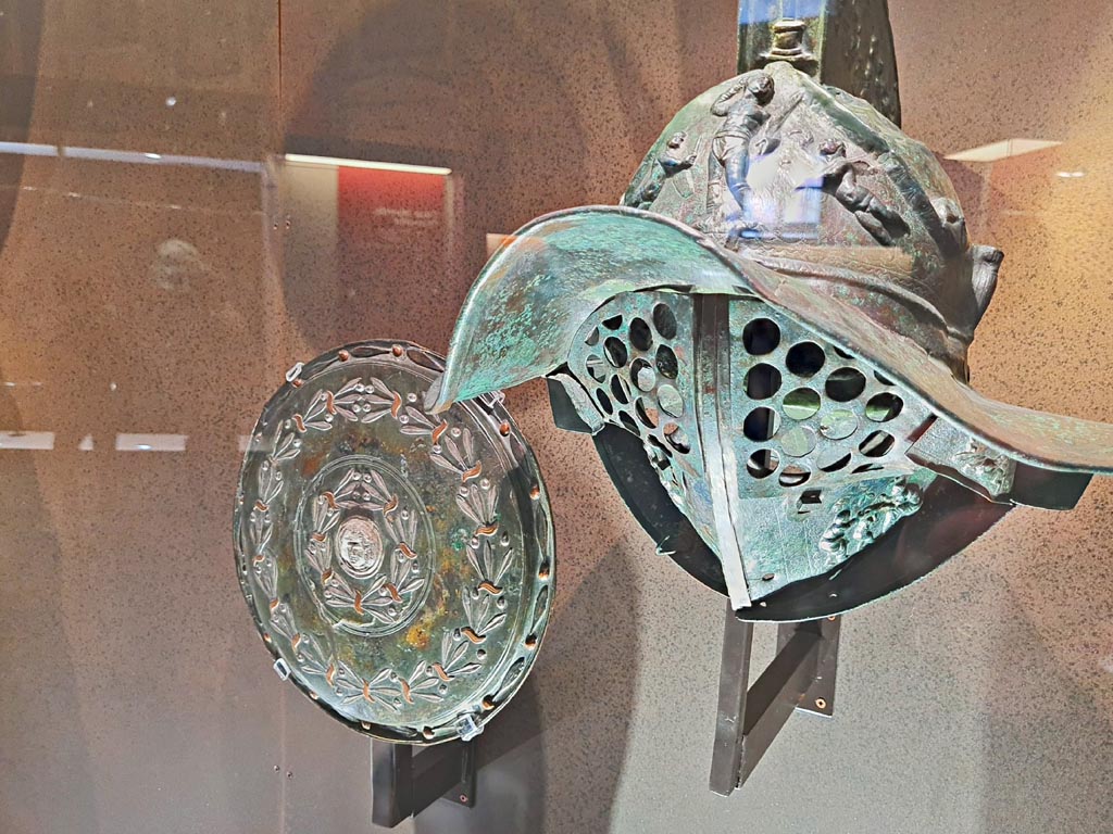 VIII.7.16 Pompeii. Photo taken May 2021, in Naples Archaeological Museum. 
On right - Murmillo gladiator’s helmet, showing Gladiator between armed cupids and a herm of Hercules and Bacchanalian scenes on crest inv. 5672.  
On left - Bronze, copper and silver shield with laurel branches and gorgeinon, inv. 5669.  Found in Gladiators Barracks, 9th January 1768.
Photo courtesy of Giuseppe Ciaramella.