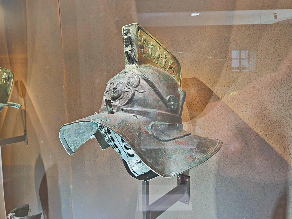 Helmet with relief of head of Medusa. Photo taken May 2021, courtesy of Giuseppe Ciaramella.
Murmillo gladiators carried heavy equipment, including a broadsword and a large, rectangular shield. 
The relief on the helmet’s crest depicts griffins being offered drinks by erotes (cupids). 
Feathers could be inserted into tubes attached to the sides of the crest. 
The head of Medusa, a mythological monster with snakes for hair, can be seen above the visor. 
Above this is Mars Ultor, the god of war, in full armour.