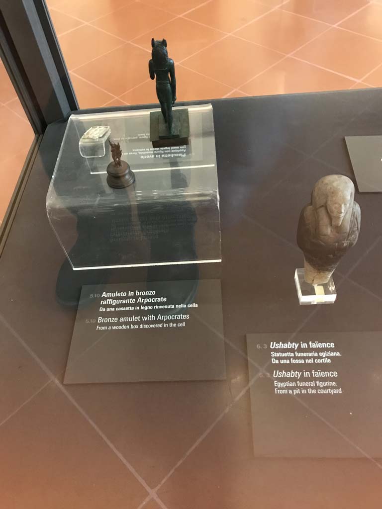 VIII.7.28 Pompeii. April 2019. Bronze amulet with Harpocrates, on left, which was found in a wooden box in the cella.  
Egyptian funeral figurine found in a pit in the courtyard, on right. 
Now in Naples Archaeological Museum. Photo courtesy of Rick Bauer.