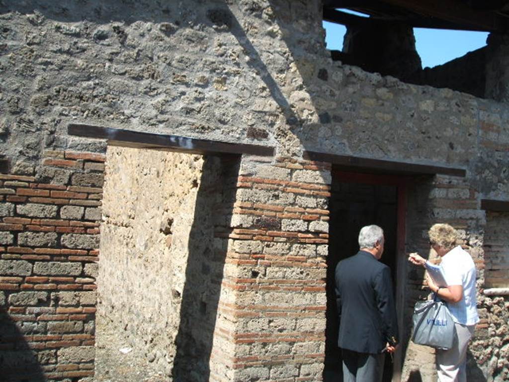 IX.5.16 Pompeii. May 2005. West side of atrium a’, on the left, doorway to kitchen and latrine e’.
On the right, doorway to cubiculum f’.
According to Boyce, on the south wall of the kitchen was a large rectangular panel of white stucco.
On it were painted two yellow serpents with red crest and beards. 
These were on either side of a tile embedded and projecting from the wall.
A fire was painted on the wall above the tile.
The background was painted with plants, and across the top were two painted garlands.
On each of the garlands was a painted bird.
See Boyce, G. K., 1937. Corpus of the Lararia of Pompeii. Rome: MAAR 14.  (p. 86, no. 425)
In IX.5.16, the first place on the left of the atrium was searched again, and found were several iron nails, some fish bones, and in terracotta: two containers for oil, a vase in fragments, two amphorae, two pot lids, and a broken bowl containing ash.
See Notizie degli Scavi di Antichità, 1885, (pp. 162-4).