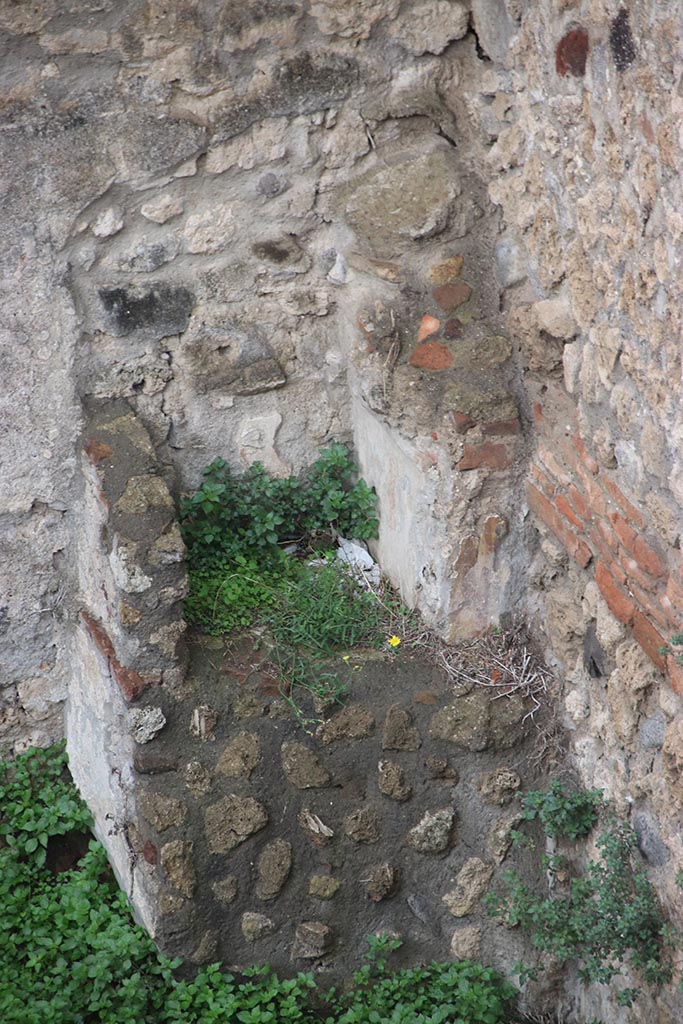 IX.7.12, 14 or 16 (?) Pompeii. October 2024. 
Garden area “e”, detail of lararium in north-west corner. Photo courtesy of Klaus Heese.
According to Boyce –
Against the west wall of the garden stands an aedicula, built upon a square masonry base (0.92 square, h.0.70).
The sides of the base are decorated with green plants painted on the white ground.
Above the base, walls enclose a niche (h.0.42, w.0.52, d.0.62).
In front of the structure stands a small altar (0.24 by 0.23, h.0.80).
The roof (no longer preserved) originally extended out over the altar as well as the niche, and the front of it was adorned with a pediment.
It was supported by two columns, the marks of the bases of which are still to be seen near the corners of the base.
On the walls within the niche is the lararium painting.
On the back wall to the right of an altar, stands the Genius pouring a libation upon it.
To the left of the same altar is a serpent, the head of which was done in stucco relief.
On each of the side walls are painted three figures: from back to front – a plant, a Lar, a man in a white garment holding in his left hand an object resembling a black cup, with a patera in his right hand.
He quotes references – Not. Scavi, 1881, 92-93; Bull. Inst., 1883, 146.
In Not. Scavi, it is described as House, only partly excavated – “and corresponds to, after the sixth doorway, west side, counting from the north-west”.
In Bull. Inst, it is described as “West side, first entrance counting from the south, garden in part excavated.”
See Boyce G. K., 1937. Corpus of the Lararia of Pompeii. Rome: MAAR 14. (p.89, no.445).