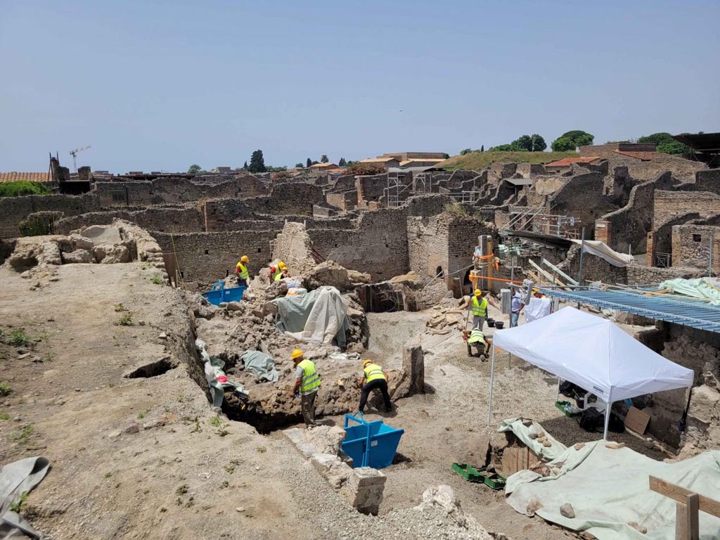 IIX.10.1 Pompeii. June 2023. View west across the house excavations.
The partially visible structures already suggested the presence of a large atrium with the canonical succession of rooms on the eastern side and, on the opposite side, the entrance to the production sector of the oven. The atrium was freed from the resulting material of the nineteenth-century excavations revealing the collapse of the compluvium roof within the layer of white pumice and a residual portion of the volcanic layers from flow (cinerites) in the southern sector. This sector still partially encumbered by the collapses preserves the frescoed tablinum (14) which occupies the entire slope with, in the south-west corner, an access corridor to a still unexplored sector. The removal of volcanic ashes has revealed in this point the surviving presence of a frescoed panel with a black background with a still life.
IX.10.1 Pompeii. Giugno 2023. Vista ovest attraverso gli scavi della casa.
Le strutture parzialmente a vista facevano già supporre la presenza di un ampio atrio con la canonica successione degli ambienti sul lato orientale e, sul lato opposto, l’ingresso al settore produttivo del forno. L’atrio è stato liberato dal materiale di risulta degli scavi ottocenteschi rivelando il crollo delle coperture compluviate all’interno dello strato di pomici bianche e una porzione residuale degli strati vulcanici da flusso (cineriti) nel settore meridionale. Questo settore ancora in parte ingombro dai crolli conserva il tablino affrescato (14) che occupa l’intero versante con, nell’angolo sud-ovest, un corridoio di accesso ad un settore ancora non esplorato. La rimozione delle ceneri vulcaniche ha rivelato in questo punto la presenza superstite di un pannello affrescato a fondo nero con natura morta.
Photograph © Parco Archeologico di Pompei.
Vedi/See Una natura morta con xenia dallo scavo della casa IX 10,1 a Pompei. E-journal 3, Scavi di Pompeii, 27.6.2023, p. 2-3. E-journal on PAP web site