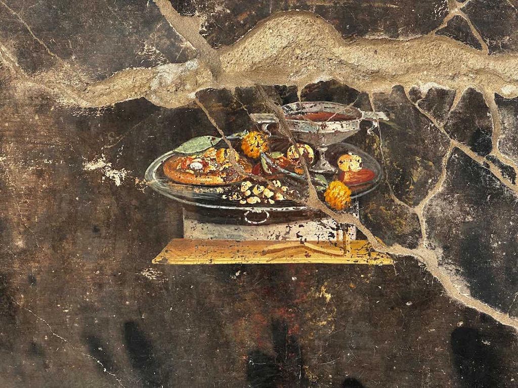 IX.10.1 Pompeii. June 2023. Atrium 2 west wall. It looks like a pizza?
Whilst it looks like a pizza, this image from a Pompeian painting from 2000 years ago obviously can't be, since some of the most characteristic ingredients are missing, namely tomatoes and mozzarella. However, it could be a distant ancestor of the modern dish, elevated to the status of a World Heritage element in 2017 as the "traditional art of the Neapolitan pizza chef'.
As the archaeologists of the Archaeological Park of Pompeii explain, it is possible to suppose that next to a wine cup placed on a silver tray, there is depicted a flat focaccia that functions as a support for various fruits, (that can be identified as pomegranate, and maybe a date), with spices and perhaps with a type of pesto, (moretum in Latin) as indicated by yellow and ochre dots, possibly condiments. Moreover, on the same tray, dried fruits and a garland of yellow strawberry trees are present next to the dates and pomegranate.
See PAP: Pompeii: a still life discovered by the new excavations of Regio IX
IX.10.1 Pompeii. Giugno 2023. Atrio 2 parete ovest. Sembra una pizza?
Sembra una pizza, quello che si vede su un dipinto pompeiano di 2000 anni fa, ma ovviamente non lo può essere, a rigore, dato che mancavano alcuni degli ingredienti più caratteristici, ovvero pomodori e mozzarella. Tuttavia potrebbe essere un lontano antenato della pietanza moderna, elevata a patrimonio dell'umanità nel 2017 in quanto "arte tradizionale del pizzaiuolo napoletano'.
Come spiegano gli archeologi del Parco Archeologico di Pompei, si suppone che accanto a un calice di vino, posato su un vassoio di argento, sia raffigurata una focaccia di forma piatta che funge da supporto per frutti vari (individuabili un melograno e forse un dattero), condita con spezie o forse piuttosto con un tipo di pesto (moretum in latino), indicato da puntini color giallastro e ocra. Inoltre, presenti sullo stesso vassoio, frutta secca e una ghirlanda di corbezzoli gialli, accanto a datteri e melograni.
Photograph courtesy of Davide Peluso.
Vedi PAP: Pompei: emerge una natura morta dai nuovi scavi della Regio IX