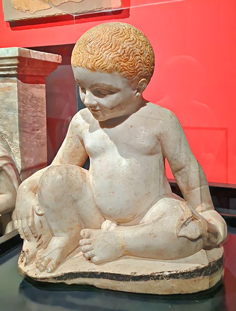 IX.12.9 Pompeii. March 2024. Fountain statuette of a boy and dolphin. Marble 1st century AD.
(Statuetta di fontana con bambino e delfino. Marmo, l sec. D.C.)
Provenanced from this house according to the information card at the exhibition entitled -
“L’altra Pompei, vite comuni all’ombra del Vesuvio”.  Photo courtesy of Giuseppe Ciaramella.
According to an information card in the house –
“The garden was also decorated with a fountain which was crowned with a statue of a young boy, on which we can identify traces of original colours.”