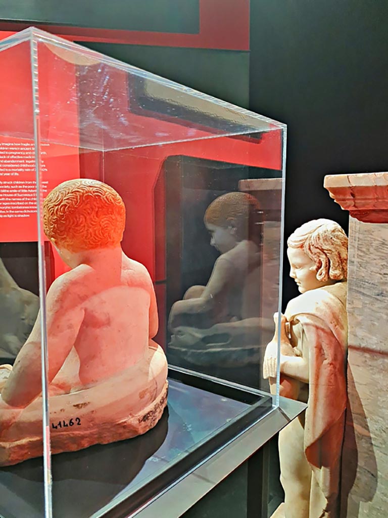 IX.12.9 Pompeii. March 2024. Fountain statuette of a boy and dolphin. Marble 1st century AD.
(Statuetta di fontana con bambino e delfino. Marmo, l sec. D.C.)
Provenanced from this house according to the information card at the exhibition entitled -
“L’altra Pompei, vite comuni all’ombra del Vesuvio”.  Photo courtesy of Giuseppe Ciaramella.
According to an information card in the house –
“The garden was also decorated with a fountain which was crowned with a statue of a young boy, on which we can identify traces of original colours.”