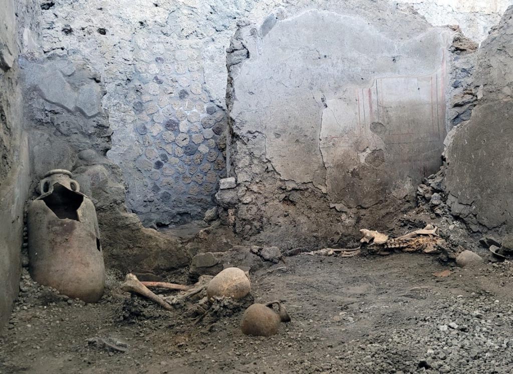 IX.12.B/IX.12.9 Pompeii. May 2023. 
Room “a”, south end of utility room with the two skeletons and an upright amphora in the corner.
They were found lying on one side in a utility room, probably not in use at the time due to repairs or renovation work under way in the house, where they had taken refuge in search of protection.
They were probably two male individuals at least 55 years old.
Estremità sud del ripostiglio con i due scheletri e un'anfora verticale nell'angolo.
Giacevano riversi su un lato, in un ambiente di servizio, al tempo in dismissione per probabili interventi di riparazioni o ristrutturazione in corso nella casa, nel quale si erano rifugiati in cerca di protezione.
Si trattava probabilmente di due individui di sesso maschile di almeno 55 anni.
Photograph © Parco Archeologico di Pompei.