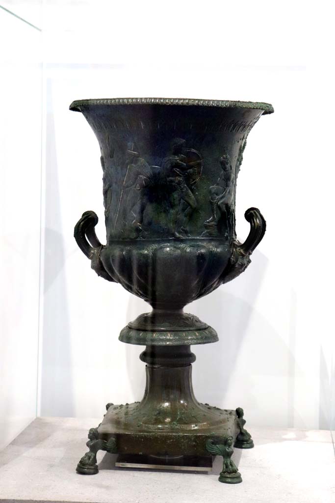 IX.13.1-3, Pompeii. February 2021. 
Bronze krater with scenes in relief, found in triclinium, on display in Antiquarium, VIII.1.4.
Photo courtesy of Fabien Bièvre-Perrin (CC BY-NC-SA).
According to the description card in the Antiquarium -
The bronze krater was decorated with eight armed male figures, six standing and two sitting. 
One of the seated figures is tying his shoelaces.
A young man stands in front of him holding a shield, with a bearded figure holding a staff behind.
Young and naked, the second seated figure caresses an animal with one hand and holds a sword by the hilt in the other.