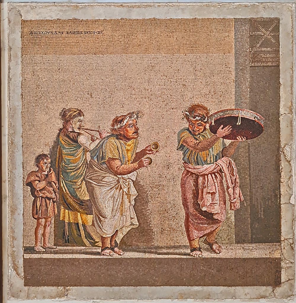 HGW06 Pompeii. Found on 8th April 1763. Ambiente 13 of Villa of Cicero. 
Mosaic of a Group of Street Musicians. It bears the signature of Dioskourides of Samos. 
Now in Naples Archaeological Museum. Inventory number 9985.
According to the information card –
Street musicians.
Mosaic emblema in opus vermiculatum made from polychrome tesserae, signed by Dioskourides of Samos.
The scene, inspired by the New Comedy, depicts three itinerant musicians with their faces covered by masks: 
a musician playing cymbals in the centre, another with a tambourine to the right and a woman with a double flute to the left. 
A boy on the left follows the procession. (Late 2nd – early Ist century BC.)
(I musici ambulanti.
Emblema pavimentale in opus vermiculatum di tessere policrome, firmato da Dioskourides di Samo.
La scena, ispirata alla Commedia Nuova, raffigura tre suonatori ambulanti col volto coperto da maschere: 
un suonatore di cembali, al centro; un altro con tamburello, a destra; una donna col doppio flauto, a sinistra.
Un fanciullo, a sinistra, segue il corteo. Fine del II – inizi del I secolo a.C.)
Photo courtesy of Giuseppe Ciaramella. September 2024.