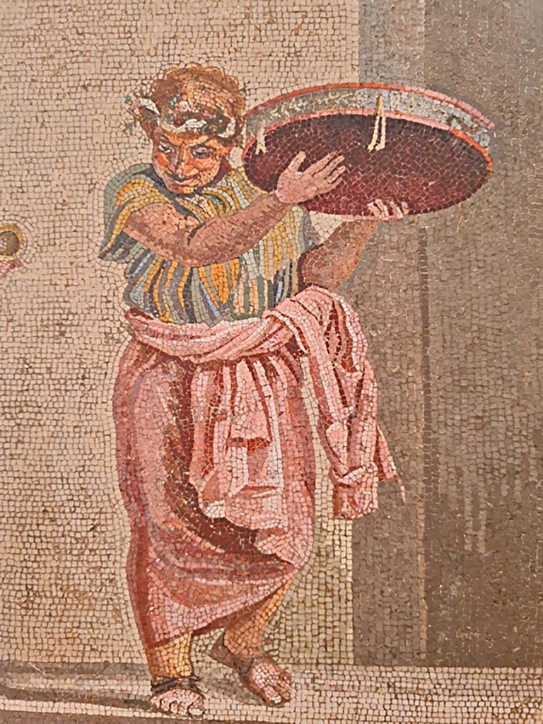 HGW06 Pompeii. Found on 8th April 1763. Ambiente 13 of Villa of Cicero. 
Detail from mosaic of a Group of Street Musicians, musician with tambourine.  
Now in Naples Archaeological Museum. Inventory number 9985.
Photo courtesy of Giuseppe Ciaramella. September 2024.