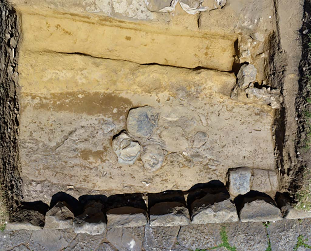 The oldest basalt road uncovered between the portico and the current paved road
Photo B. Lemaire, CJB CNRS/EFR - CC BY-NC-SA 4.0
We observed in two holes the remains of an older basalt road, lying at a lower level than the current one via dei Sepolcri, and having a slightly different orientation. 
The negative trace left by the blocks of the sidewalk shows a fairly clear difference between the ancient route and the current road.
La route en basalte la plus ancienne mise au jour entre le portique et l’actuelle voie dallée. 
Cliché B. Lemaire, CJB CNRS/EFR - CC BY-NC-SA 4.0
Nous avons pu observer dans deux sondages les restes d’une voie en basalte, plus ancienne, se trouvant à un niveau plus bas que l’actuelle via dei Sepolcri, et ayant une orientation légèrement différente. 
La trace en négatif laissée par des blocs du trottoir permet d’observer une différence assez claire entre le tracé antique et l’actuelle voie.
Pompéi : Le secteur de la Porte d’Herculanum à Pompéi : Entre espace funéraire et commercial : organisation, gestion et transformations d’une zone suburbaine 
https://centrejeanberard.cnrs.fr/spip.php?article107&lang=fr