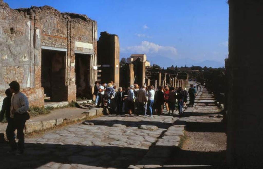Via dell’ Abbondanza, Pompeii. February 1988.
Looking east, with VII.9.68/67 doorways on left, behind fountain surrounded by crowd of tourists.
Photo by Joachime Méric courtesy of Jean-Jacques Méric.