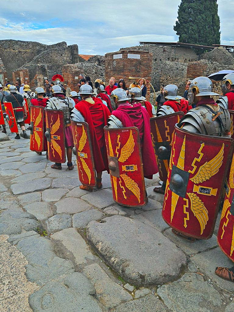 Via dell’Abbondanza, south side, Pompeii. 28th September 2024. 
Legionaries with shields with the Cornicen at the side with the trumpet.
Looking south-east towards VIII.4.1 on east side of Via dei Teatri, during “Ludi Pompeiani” event. 
Photo courtesy of Giuseppe Ciaramella.