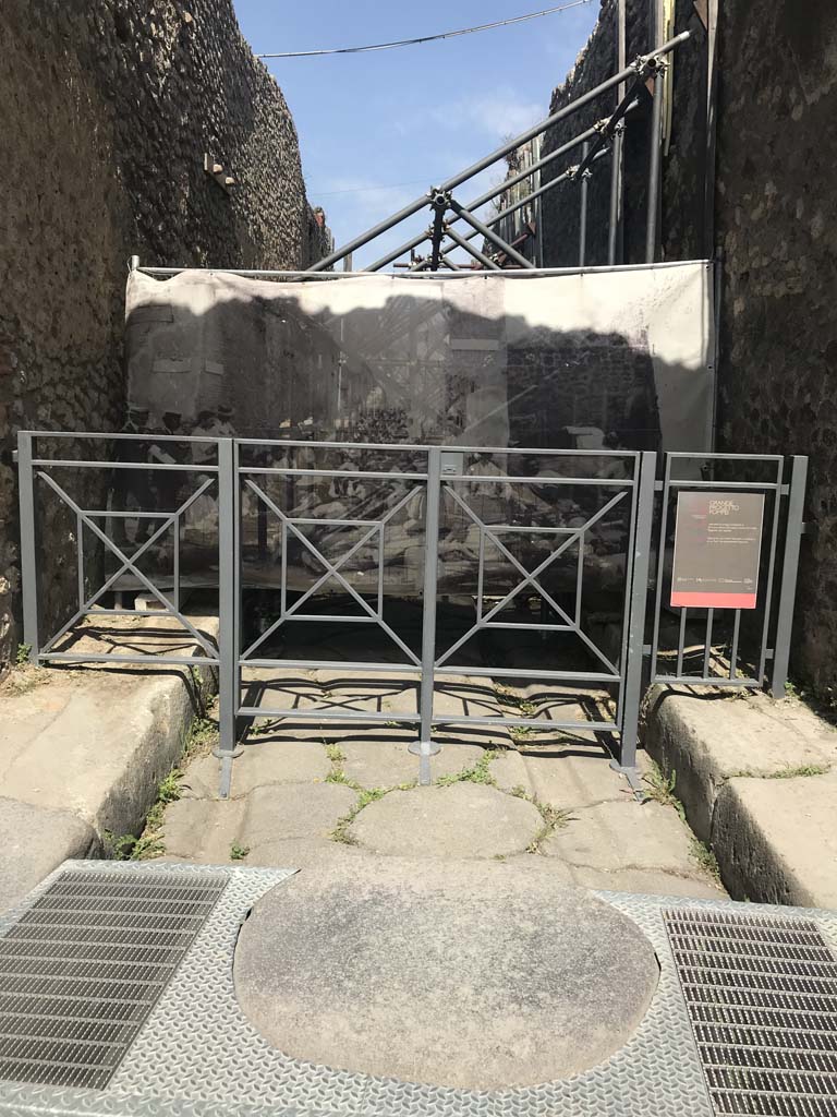 Vicolo di Tesmo between IX.1 and IX.7. April 2019. Looking north from Via dell’Abbondanza.
The screen hiding the vicolo shows an enlargement of a photo of the “first workers resting in the shade of the vicolo and posing for their photo.”
See Spinazzola, V. (1953). Pompei, alla luce degli Scavi Nuovi di Via dell’Abbondanza, 1910-1923. (p.31, fig.30)
Note the modernised accessible stepping-stone in the above photo.  Photo courtesy of Rick Bauer.