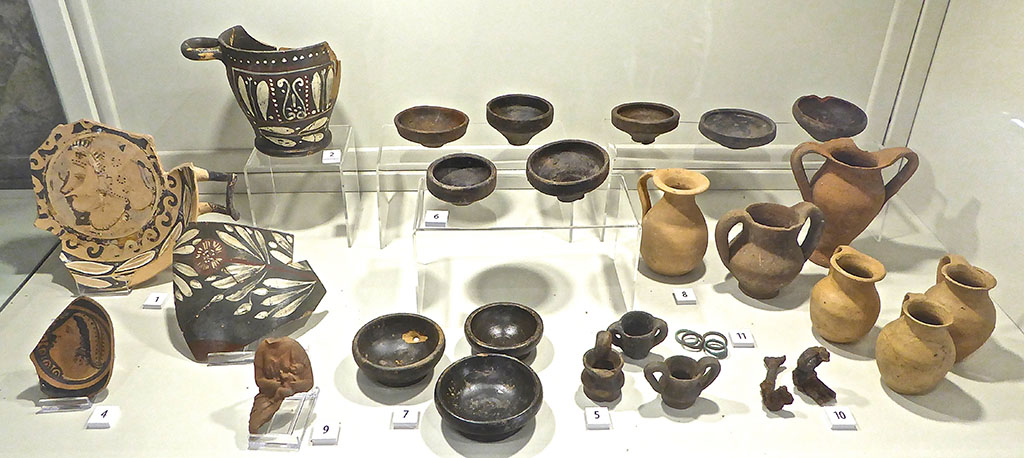 Santuario extraurbano del Fondo Iozzino. June 2017. Finds from site. Now in PAP deposits.
Photo courtesy of Michael Binns.
The exhibition information card identifies these as:
1:   Red figure kylix with woman’s head, 4th century BC
2:   Campanian painted skyphos, second half of 4th century BC
3:   Rim of painted Campanian bowl, second half of 4th century BC
4:   Base of Campanian bowl with female head, second half of 4th century BC
5:   Miniature kraters, 4th to 2nd century BC
6:   Miniature plates, 3rd to 2nd century BC
7:   Black gloss cups, 3rd century BC
8:   Coarse ware beakers, 4th to 2nd century BC
9:   Terracotta figurine of woman nursing a child, 4th to 3rd century BC
10:  Votive iron keys, Hellenistic period
11:  Bronze rings, Hellenistic period