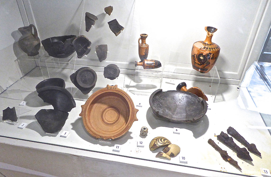 Santuario extraurbano del Fondo Iozzino. June 2017. More finds from site. Now in PAP deposits.
Photo courtesy of Michael Binns.
The exhibition information card identifies these as:
1:   Campanian bucchero kantharos with Etruscan inscription, “mi marmarces tetanas” = “of Marmarce Tetana”, second half of 6th century BC
2:   Campanian bucchero kantharos with Etruscan inscription, second half of 6th century BC
3:   Campanian bucchero bowl with graffito bird, second quarter of 6th to 1st quarter of 5th century BC
4:   Bucchero chalice with fan motifs, end of 7th to 1st quarter 6th century BC
5:   Bucchero olpe, second half of 6th to 1st quarter of 5th century BC
6:   Campanian bucchero bowl, second half of 6th century BC
7:   Campanian bucchero kantharos, second half of 6th century BC
8:   Attic black figure lekythoi with palmette decoration, circa 480 BC
9:   Attic black figure lekythoi with hoplites, of 6th to beginning of 5th century BC
10:  Corinthian aryballos decorated with hoplites, 580 to 560 BC
11:  Bowl with painted decoration, archaic period
12:  Faience bead, archaic period
13:  Iron arrow heads, 6th century BC
14:  Sherds of bucchero kantharoi and cups with graffiti, 6th century BC